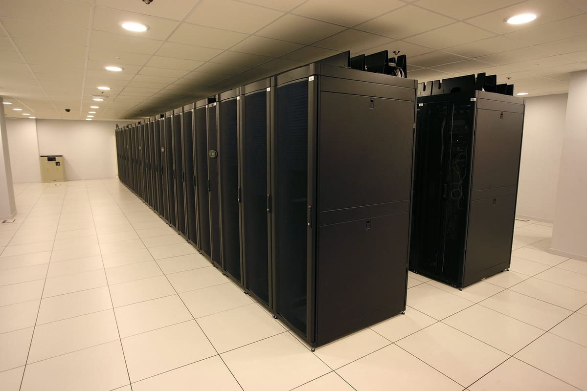Pasillo de una sala de CPD con una fila larga de racks negros cerrados sobre suelo técnico blanco.