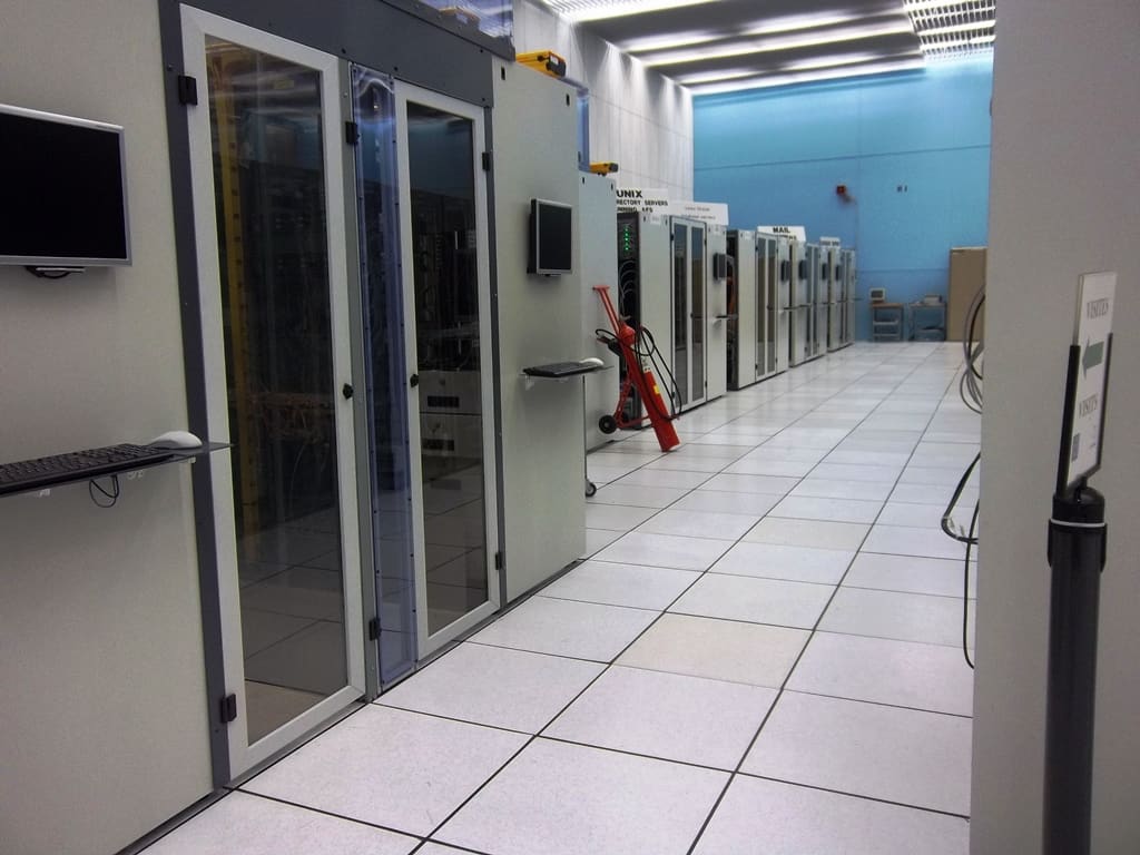 Pasillo de armarios rack con puertas de cristal, monitores KVM y carro extintor rojo, cartel de visitas