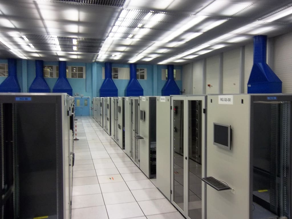 Pasillo de armarios rack grises con monitor y teclado KVM y grandes conductos azules descolgados del techo