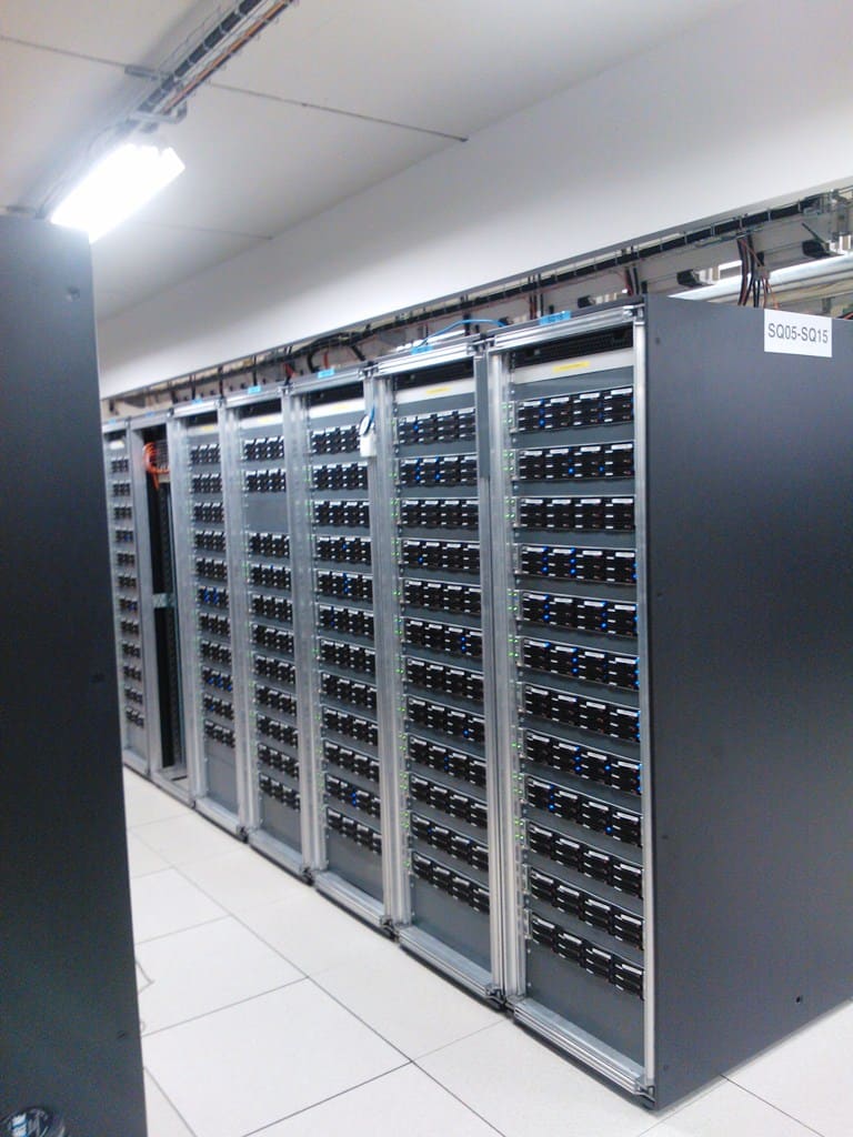 Fila de racks abiertos con bastidor de aluminio repletos de nodos de cálculo, etiqueta SQ05-SQ15 en el extremo del pasillo.