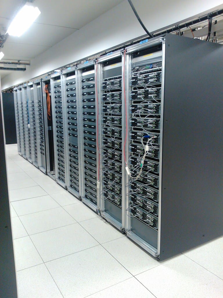 Pasillo de racks abiertos con servidores densamente apilados y cableado naranja y rojo sobre suelo técnico.