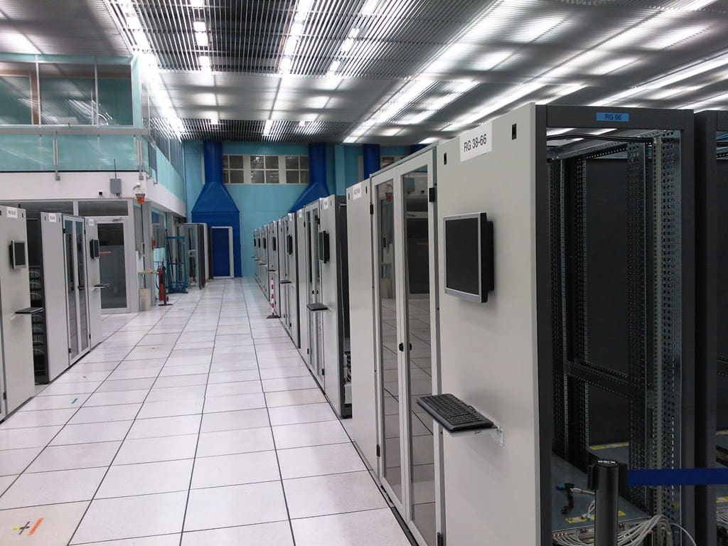 Largo pasillo de armarios de rack blancos; uno abierto muestra el bastidor negro vacío, con teclado y monitor en la puerta.