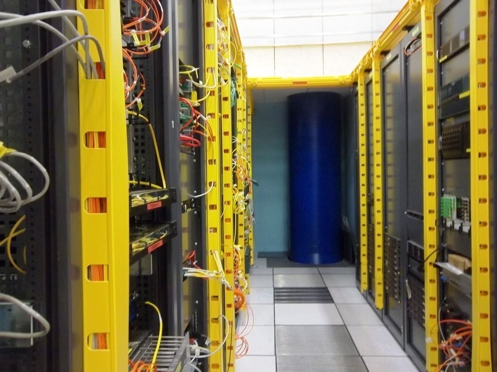 Pasillo entre racks abiertos con raíles amarillos y densa fibra naranja; columna azul al fondo del CIXP