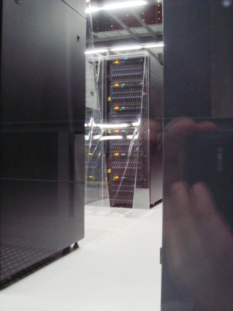 Pasillo entre armarios negros con un rack de nodos de LEDs de colores al fondo, reflejo del visitante en el cristal