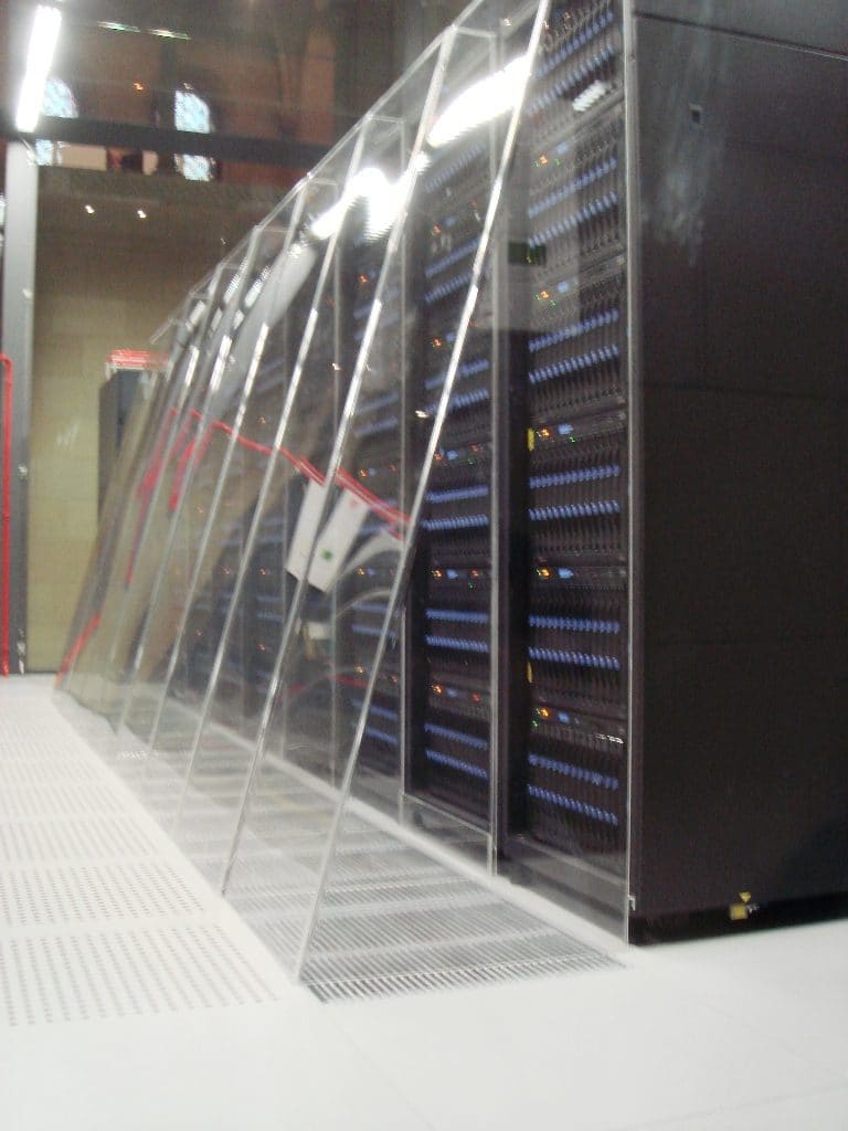 Hilera lateral de racks tras la urna de cristal inclinada, con nodos de LEDs azules sobre suelo técnico perforado